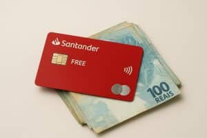 Um cartão de crédito vermelho do Santander Free repousa sobre um pequeno maço de notas de 100 reais, em fundo neutro bege claro. O cartão exibe o chip dourado, o logotipo do Santander e o símbolo de pagamento por aproximação. A imagem transmite sensação de elegância, modernidade e praticidade financeira.